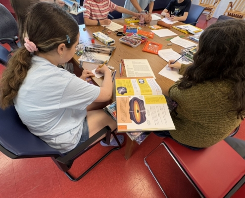 Youth sitting around a table with research books opens and colourful paper and pencils.